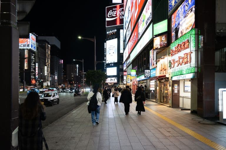 Nighttime city street scene with illuminated signs in Japanese, pedestrians walking on sidewalk, and cars on the road.