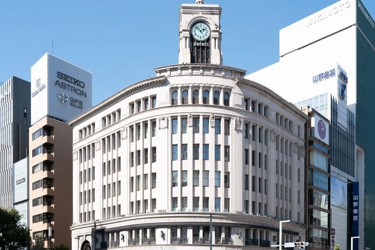 A large, historic white building with a clock tower on top, situated on a busy city street with cars, motorcycles, and pedestrians. Modern skyscrapers surround it.