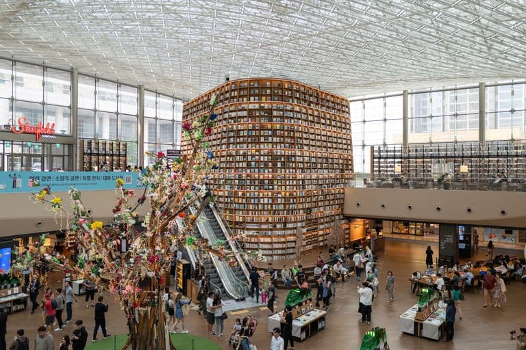 Interior of a modern library with a large circular bookshelf, a colorful decorative tree, and people browsing and reading.
