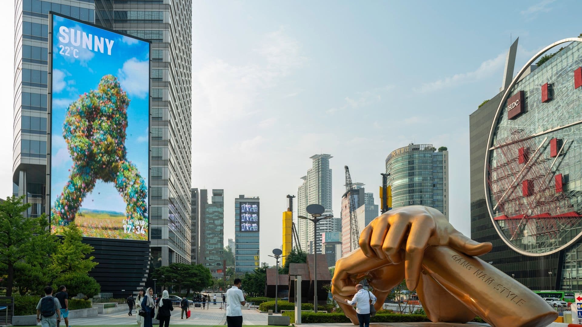 A large golden sculpture of a hand with fingers curled, situated in an urban park area surrounded by tall modern buildings, with people walking and standing nearby.