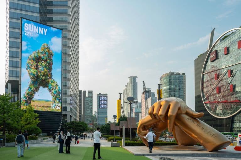 A city square featuring a large golden hand sculpture with the inscription 'GE KIM NAM STYLE', surrounded by people, skyscrapers, digital billboards, and a glass-encased building with a red logo.
