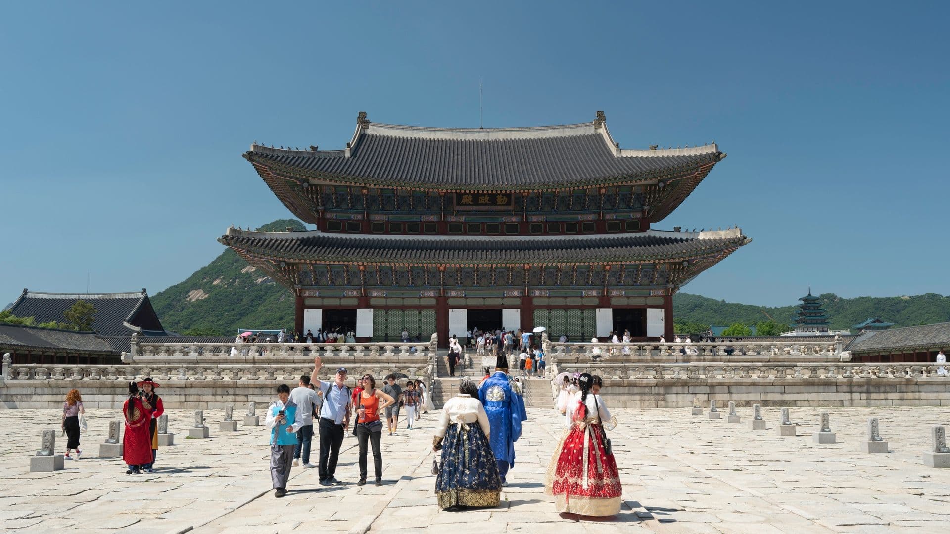 Traditional Asian temple with people in colorful traditional clothing in front, blue sky, green mountains in background.