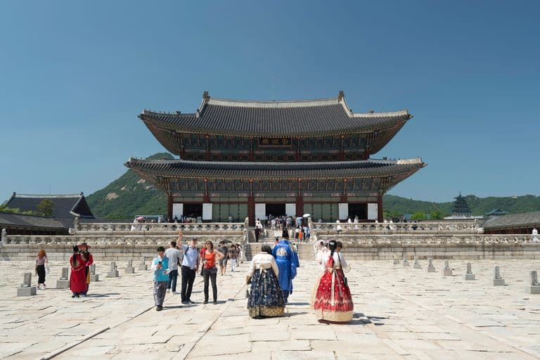 People visiting a traditional Korean palace building with a multi-tiered roof, surrounded by a stone courtyard, some dressed in traditional Korean clothing, with mountains in the background on a clear, sunny day.