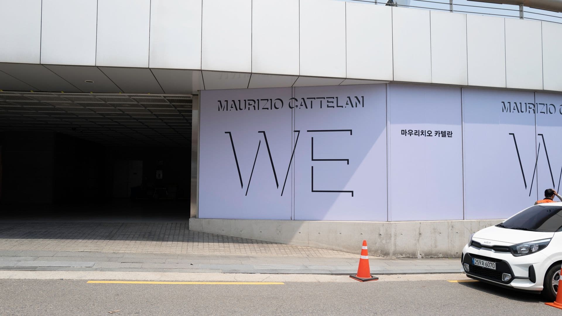 Exterior of a building with large white walls displaying "Maurizio Cattelan" and "WE" in black letters, a car parked nearby, and orange traffic cones.