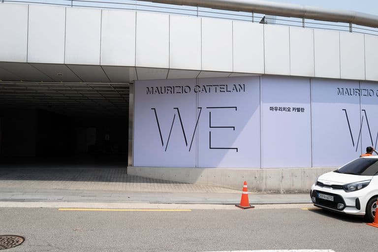 Large advertising billboard for Maurizio Cattelan's art exhibit with the word 'WE' and Korean text, located on the exterior wall of a modern building, next to a parked white car and traffic cones.