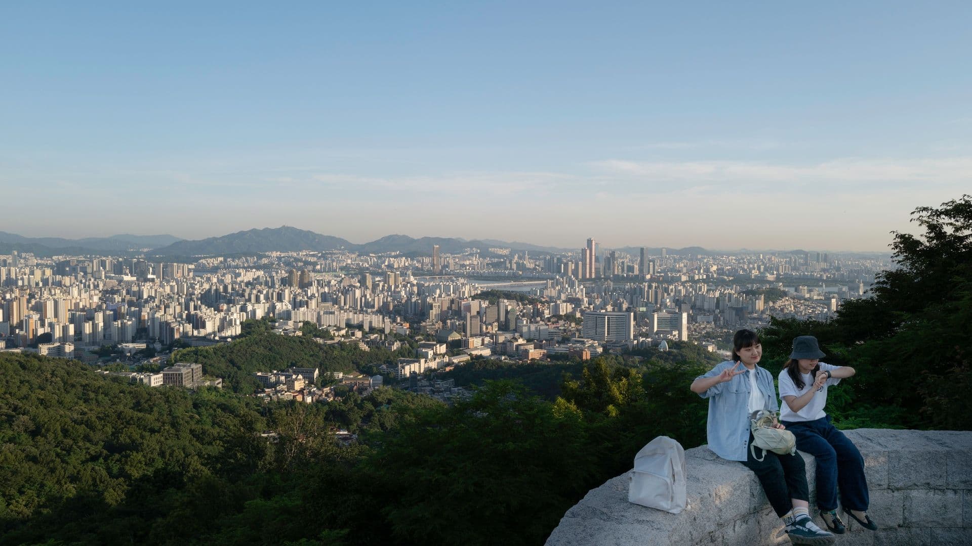 Two women sitting on a large rock ledge overlooking a city skyline with numerous high-rise buildings, and green trees in the foreground on a clear day.