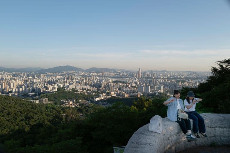 Two girls sitting on a stone ledge overlooking a cityscape with numerous high-rise buildings and mountains in the distance, taking selfies and making peace signs during daytime.