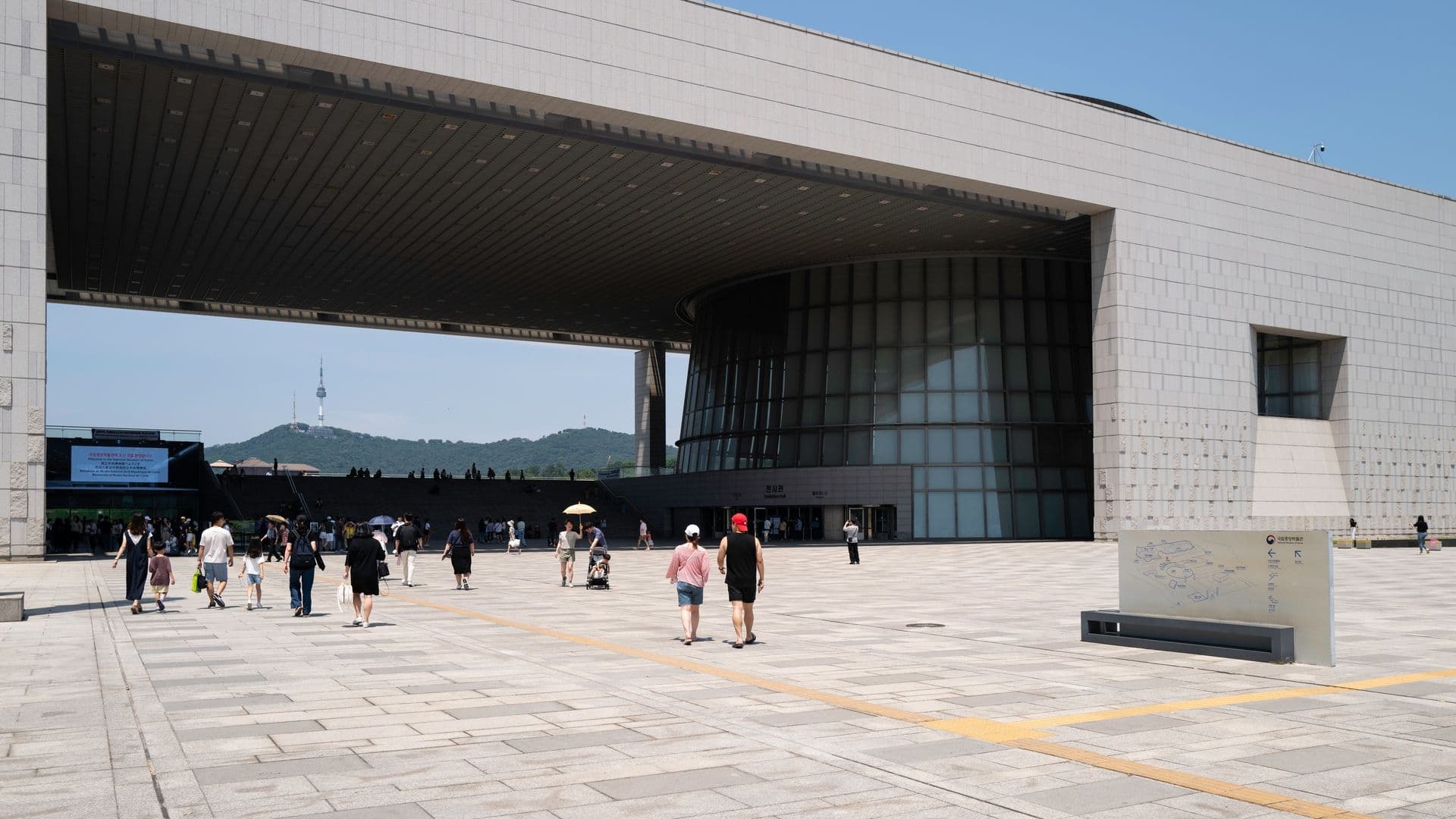 Modern public building with wide open plaza, people walking, and hills with a tower in the background under clear blue sky.