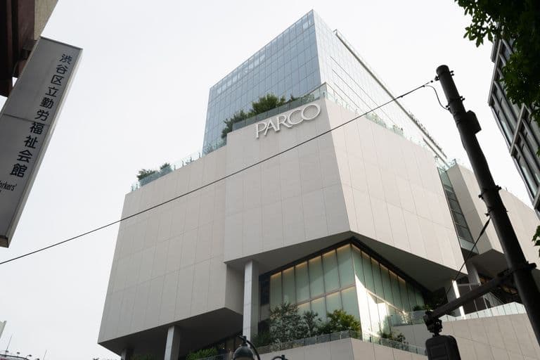 Modern multi-story building with the sign "PARCO" on it, featuring large glass windows and outdoor greenery at street level, viewed from a low angle.