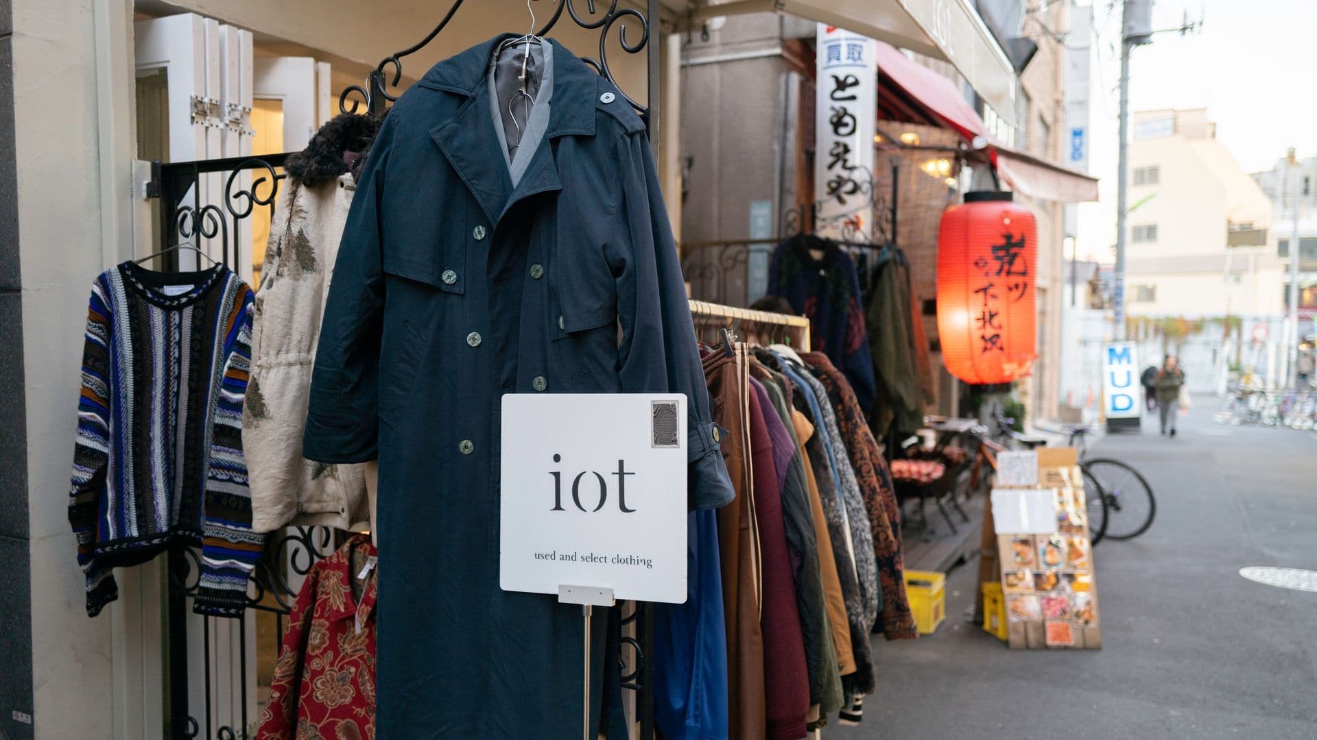 Clothing for sale outside a shop, including jackets, coats, and sweaters, with a sign reading "iot" and a red Japanese paper lantern hanging nearby.