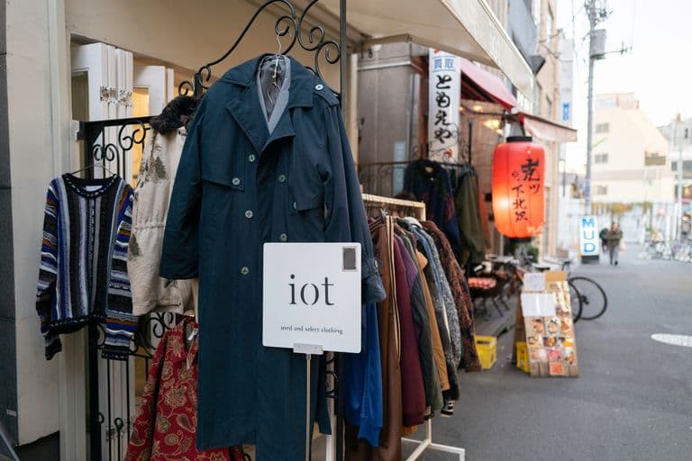 Street clothing shop with racks of used and select clothing, including a navy trench coat, a colorful patterned sweater, and skirts, with a red paper lantern hanging outside and street scenes in the background.
