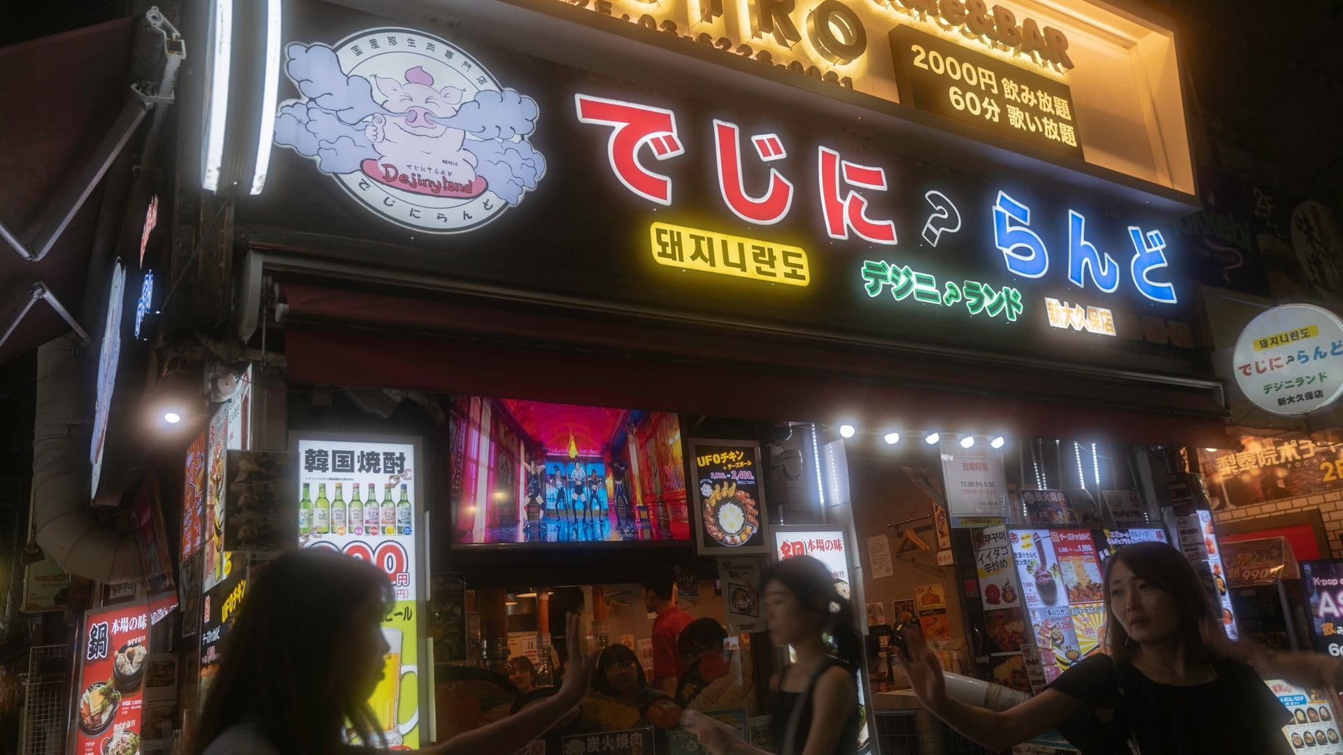 Nighttime street scene in Japan with illuminated signs, including a large sign with Japanese characters, a cartoon bear mascot, and people walking and gesturing in front of a restaurant or bar.