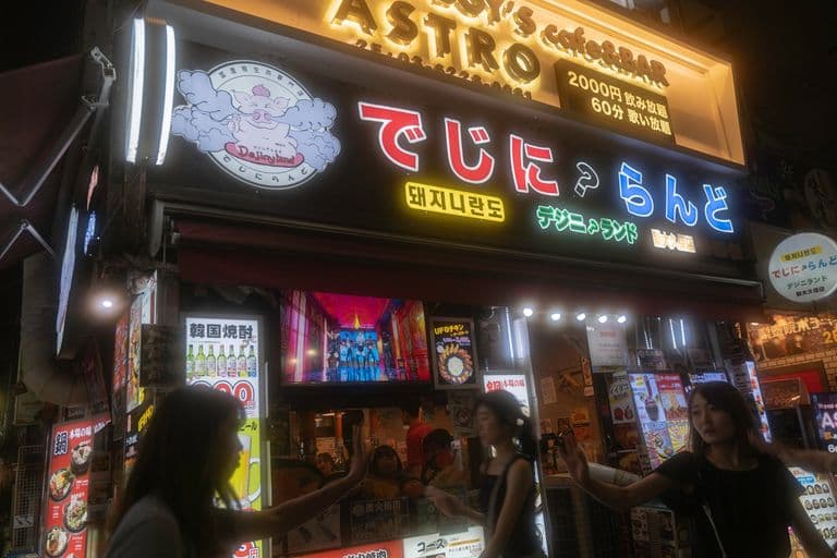 Night scene of a Japanese fast food restaurant with bright neon signs, Japanese text, and several people interacting outside.
