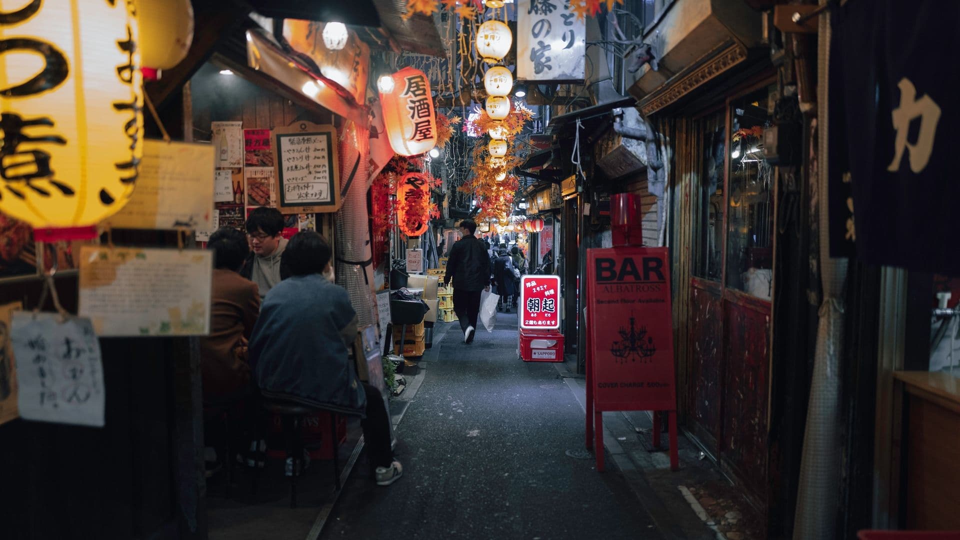 A narrow, lively alleyway in Japan decorated with hanging lanterns and colorful autumn-themed decorations, with several people sitting and walking.