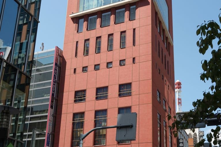 Tall pink building with large upper windows and smaller lower windows in an urban area, with people crossing the street below.