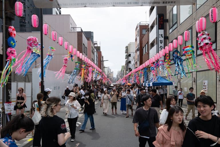 Street scene during festival with people walking, pink lanterns, and colorful paper streamers hanging overhead.