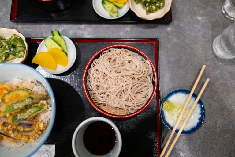 A Japanese meal featuring a bowl of cold soba noodles, a small dish of sliced cucumbers and pickled yellow radish, a bowl of rice topped with tempura vegetables, a small dish of chopped green onions and wasabi, a cup of dipping sauce, and a glass of water, arranged on black trays on a gray table.
