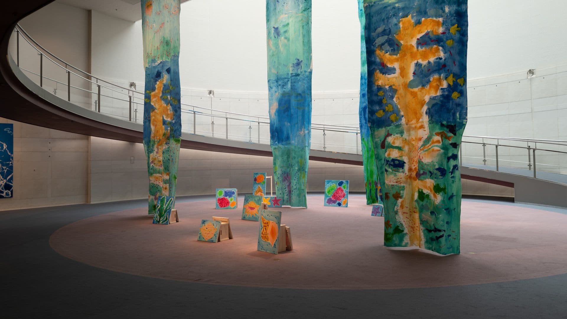 An art installation featuring large vertical fabric banners and smaller paintings, all with colorful abstract and nature-inspired designs, displayed in a spacious indoor gallery with a curved walkway and soft pink flooring.