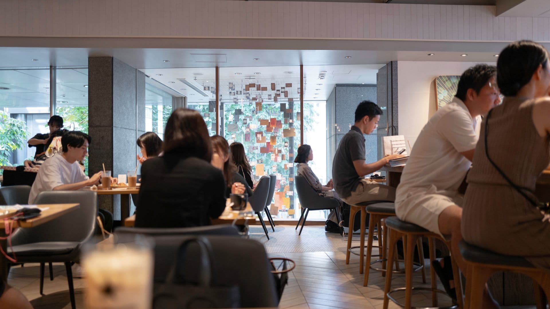 People sitting at tables and a bar in a modern restaurant or cafe with large windows and colorful art installation in the background.
