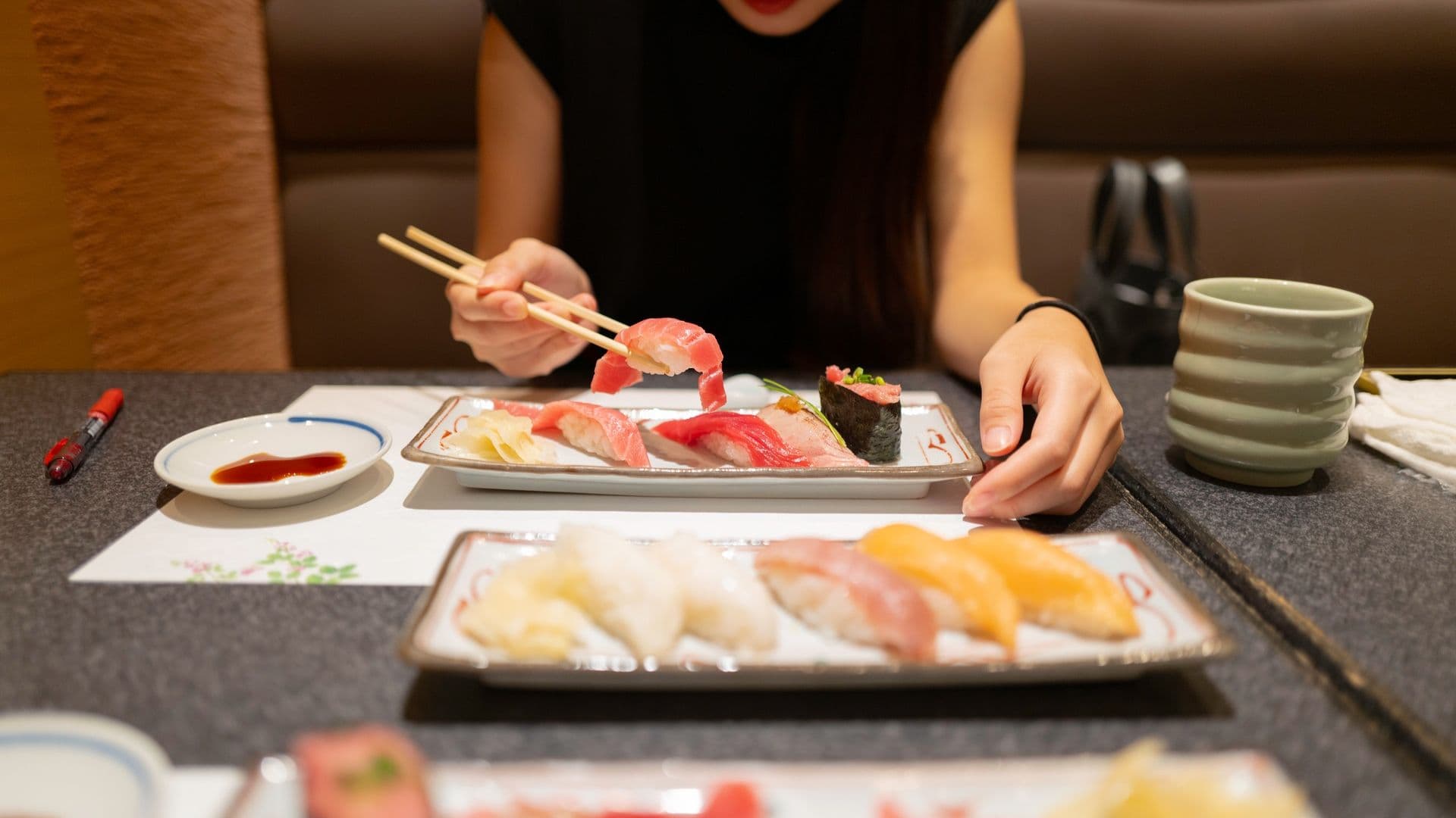 A woman sitting at a table with various plates of sushi, using chopsticks to pick up a piece of sushi, with soy sauce, a ceramic cup, and other sushi on the table.