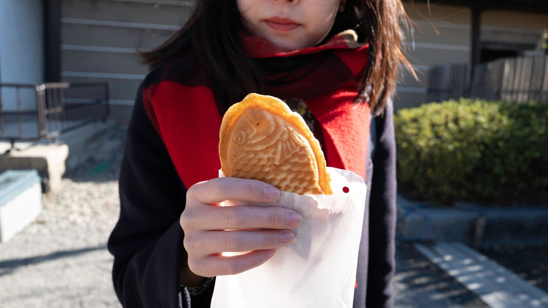 A woman holding a taiyaki fish-shaped pastry in a paper bag outdoors.