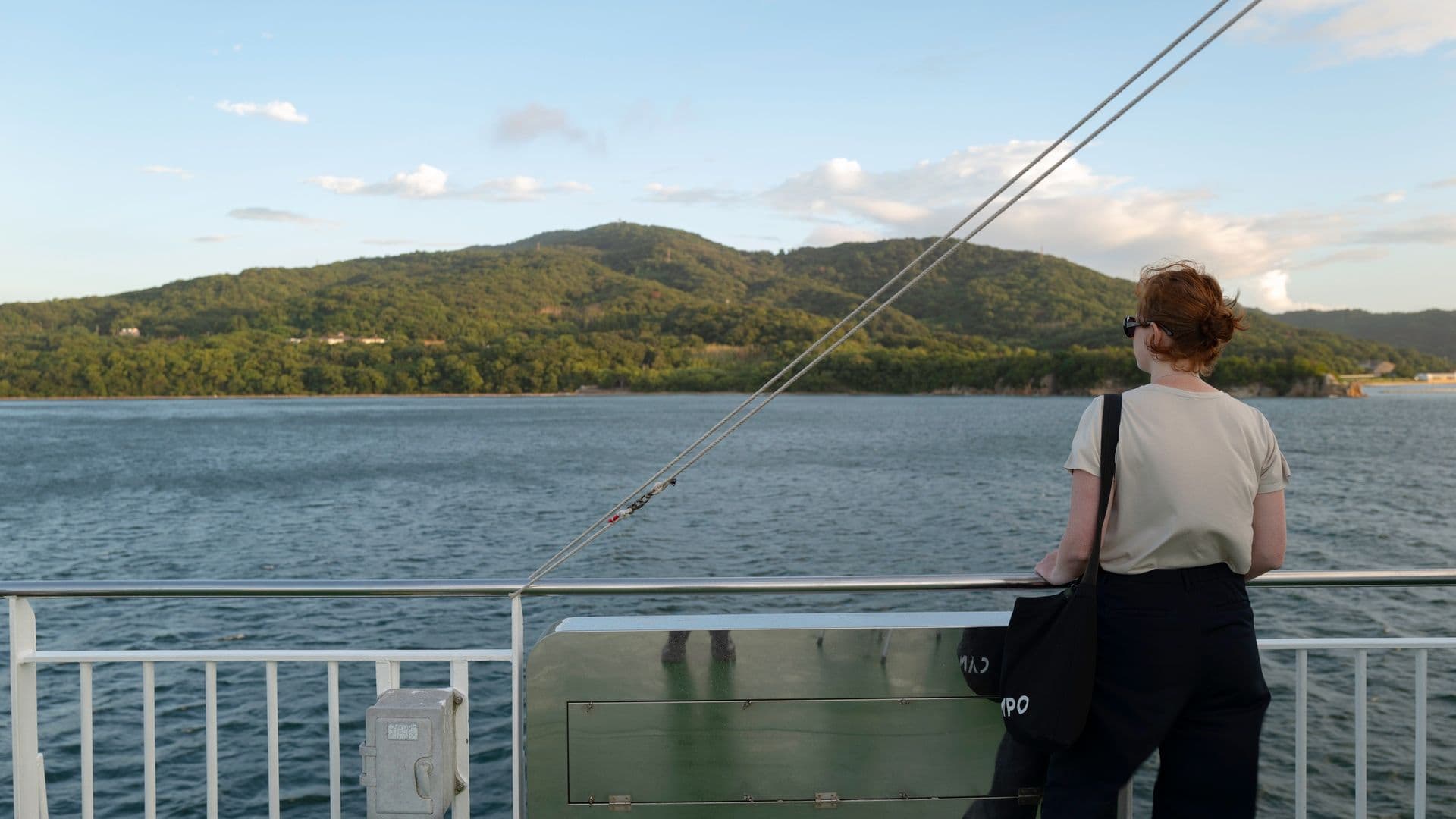 A person with short hair and sunglasses standing on a boat railing, looking at the water and distant green mountains during sunset or late afternoon.