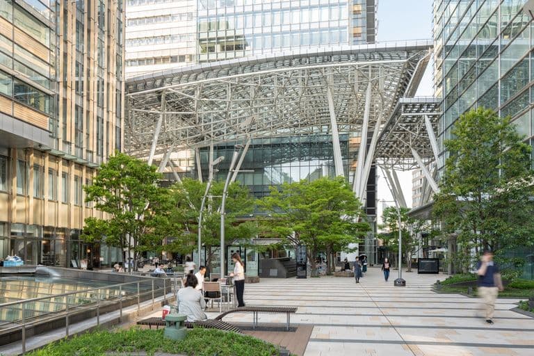 A modern urban plaza surrounded by tall glass buildings, with trees, benches, and people walking or sitting.