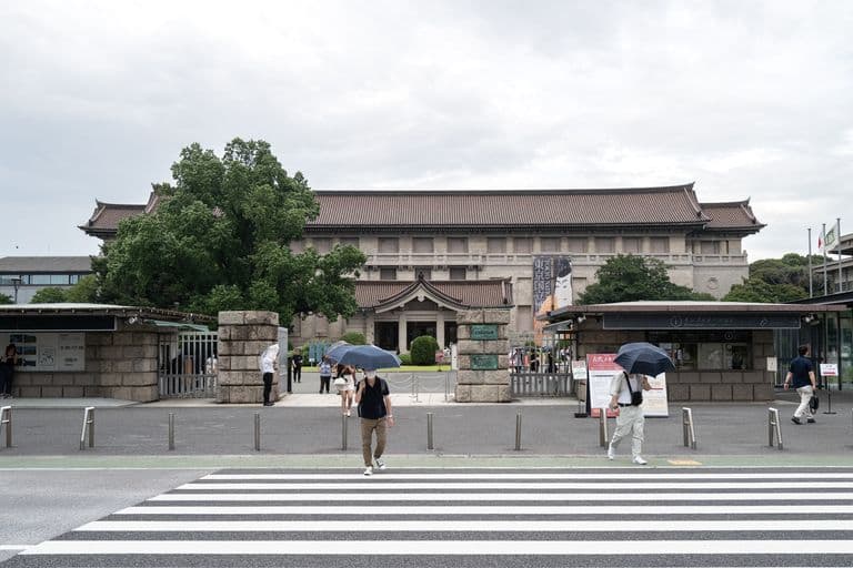 People walking with umbrellas in front of a historic building with traditional Asian architecture and green trees.