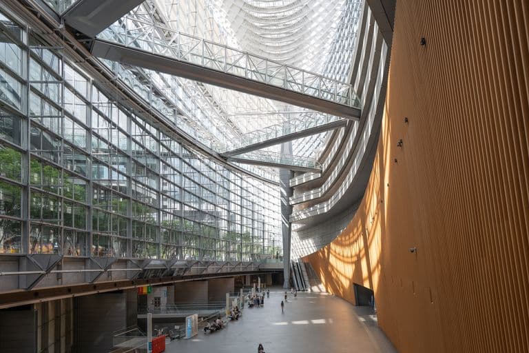 Interior of a modern building with large glass walls, high ceilings, steel beams, and a curved wooden wall, with people sitting and walking in the spacious open area.