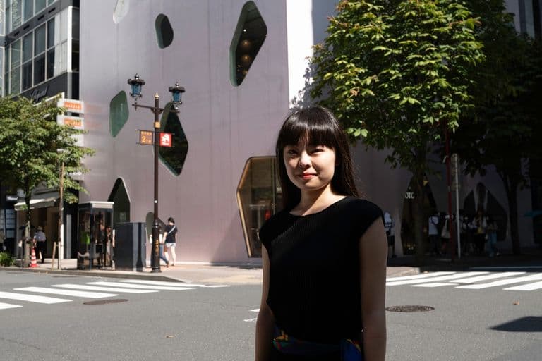 A young woman in black dress standing on city street with modern building featuring irregularly shaped windows and trees in background.