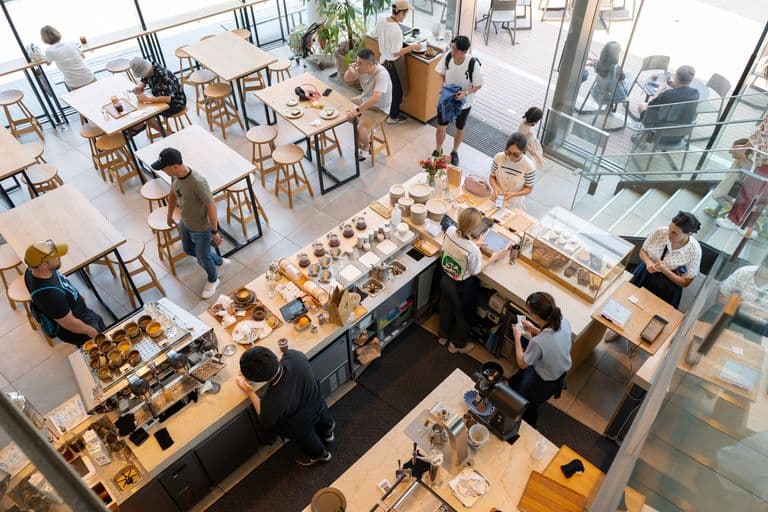 Aerial view of a modern coffee shop with customers sitting inside and outside, baristas preparing drinks, and a pastry display case.