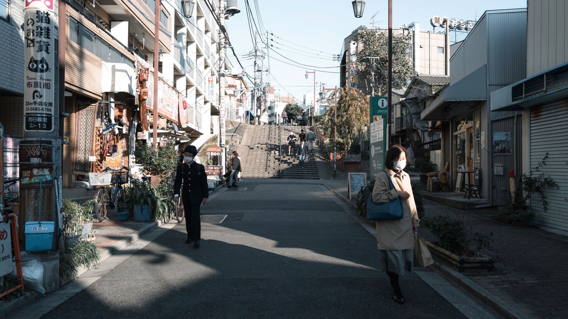 A street scene in Japan with shops, stairs leading up, and people wearing masks walking along the sidewalk.