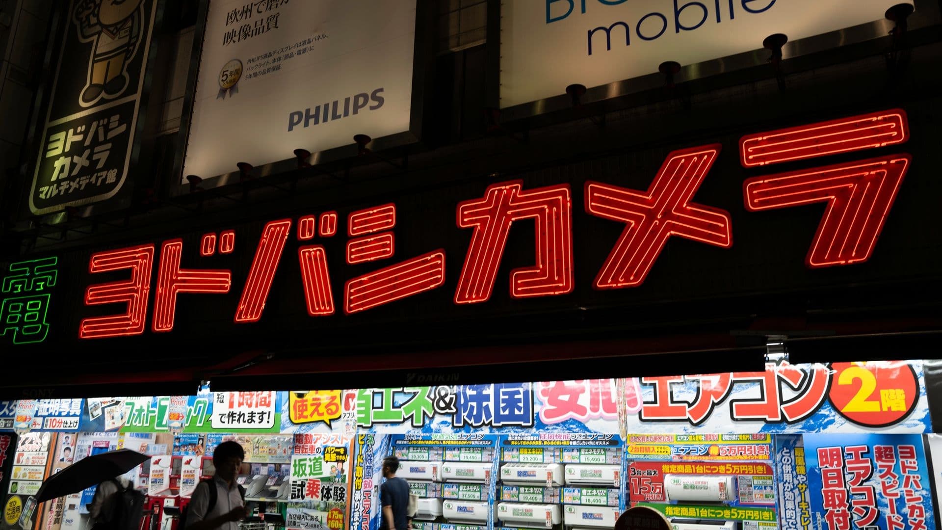 Bright neon sign in Japanese above an electronics store entrance showing various colorful advertising posters and people walking outside at night.