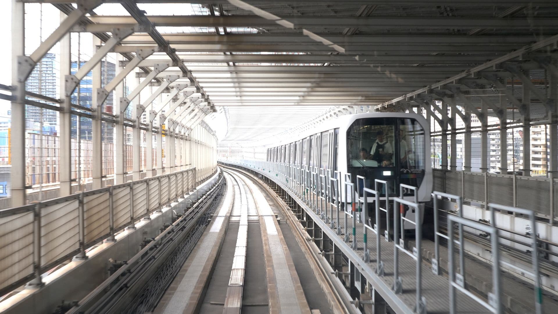 An elevated train moving through an outdoor station with a city skyline in the background.