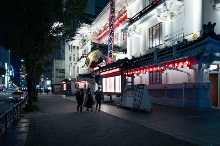 Nighttime street scene outside a large illuminated Japanese theater with red lanterns and traditional architectural details, with pedestrians walking along the sidewalk.