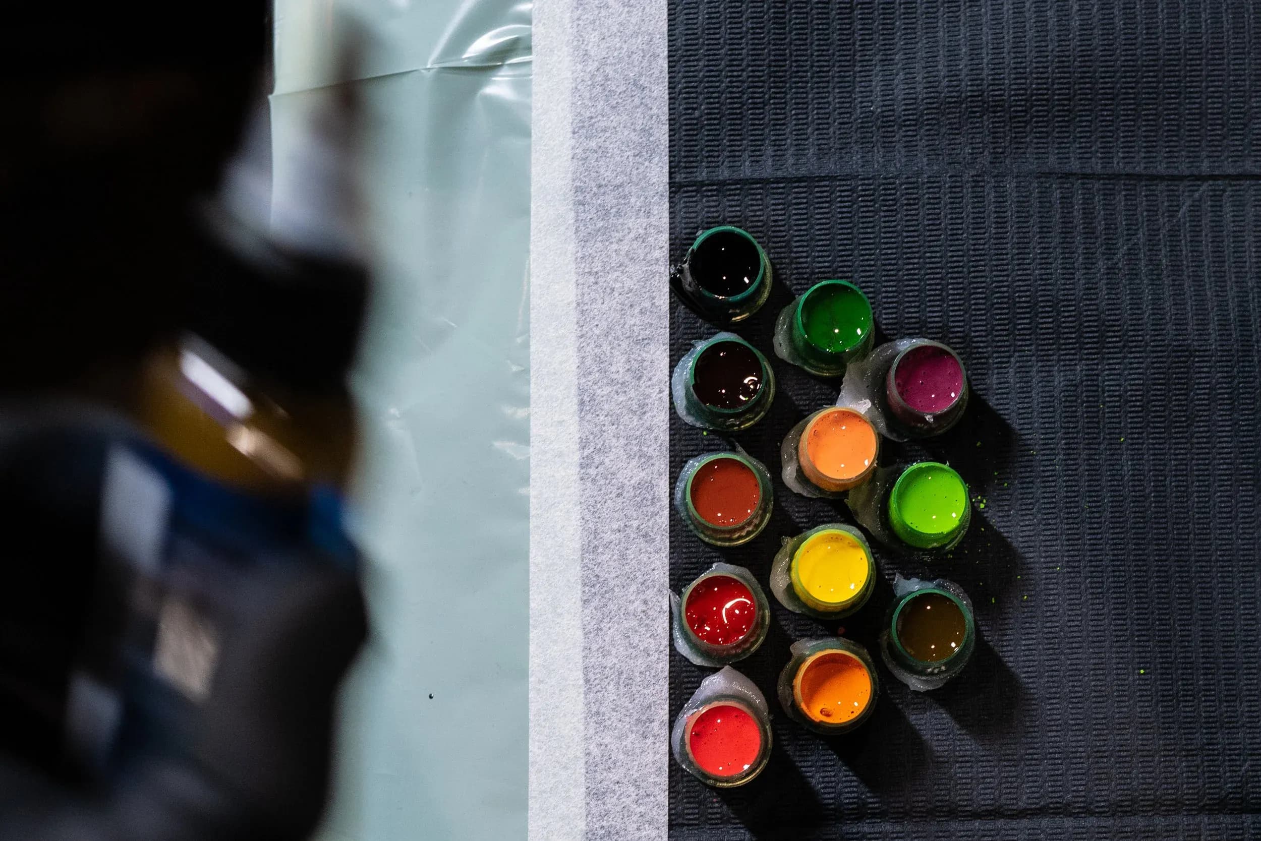 Various small cups filled with brightly colored paints on a textured black surface, accompanied by an obscured, out-of-focus bottle on the left.