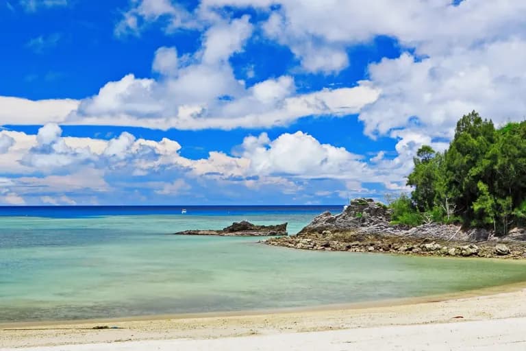 A tropical beach with calm turquoise water, rocky outcroppings, and lush green trees under a partly cloudy sky.