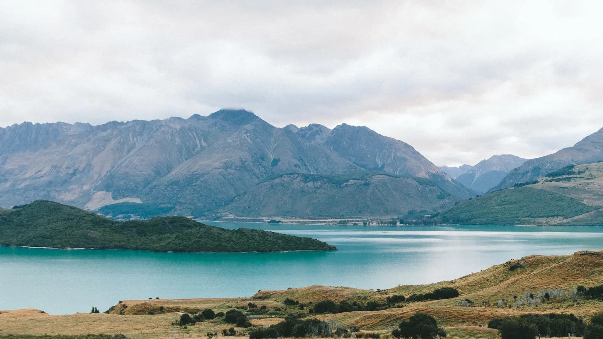 Scenic view of turquoise lake surrounded by mountains and grassy hills under a cloudy sky.