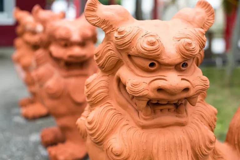 Close-up of a terracotta Chinese guardian lion statue with detailed facial features, fur, and open mouth, with other similar statues blurred in the background.