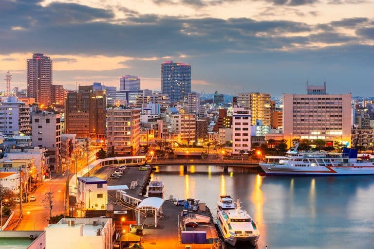City skyline at dusk with illuminated high-rise buildings, waterfront with boats, and cloudy sky.