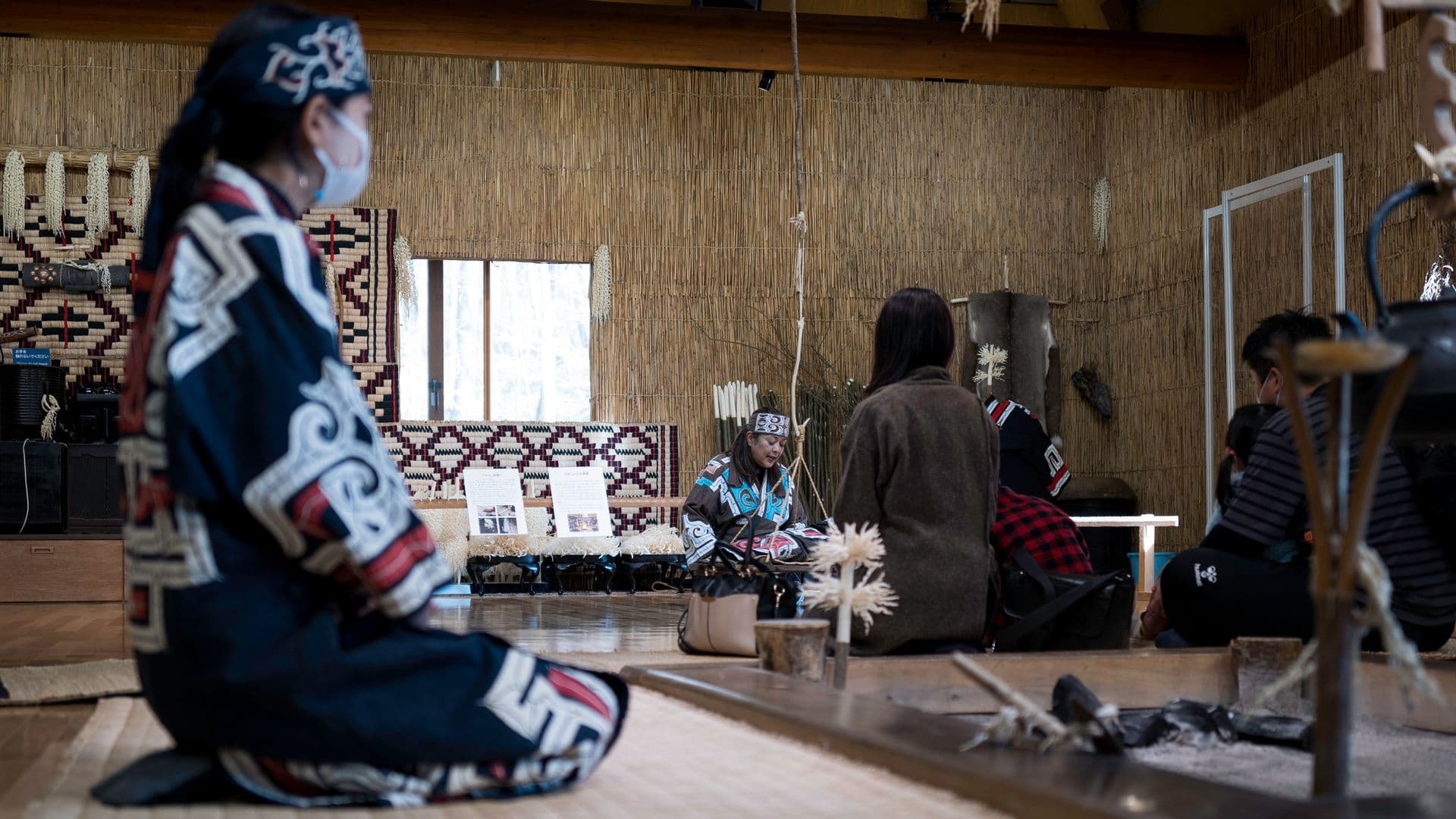 Group of people participating in a traditional indigenous ceremony, sitting on tatami mats inside a room with bamboo walls, some wearing face masks, and a woman in traditional dress leading the ceremony.