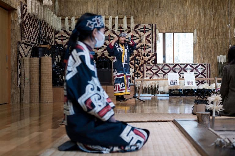 Two women dressed in traditional Indigenous clothing in a room with woven wall hangings and informational boards, one woman standing and speaking into a microphone, the other kneeling on the floor with her hands resting on her knees.