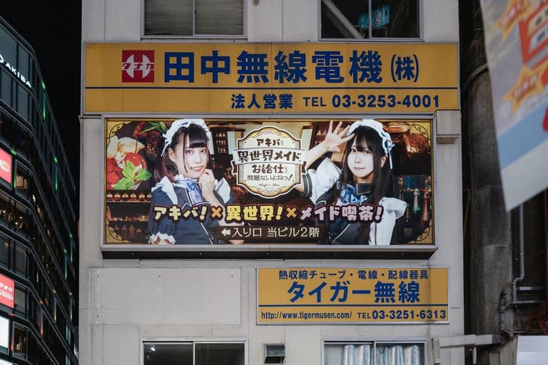 Billboard featuring two women dressed in maid costumes, promotional text in Japanese, and a background of a bar or cafe setting.
