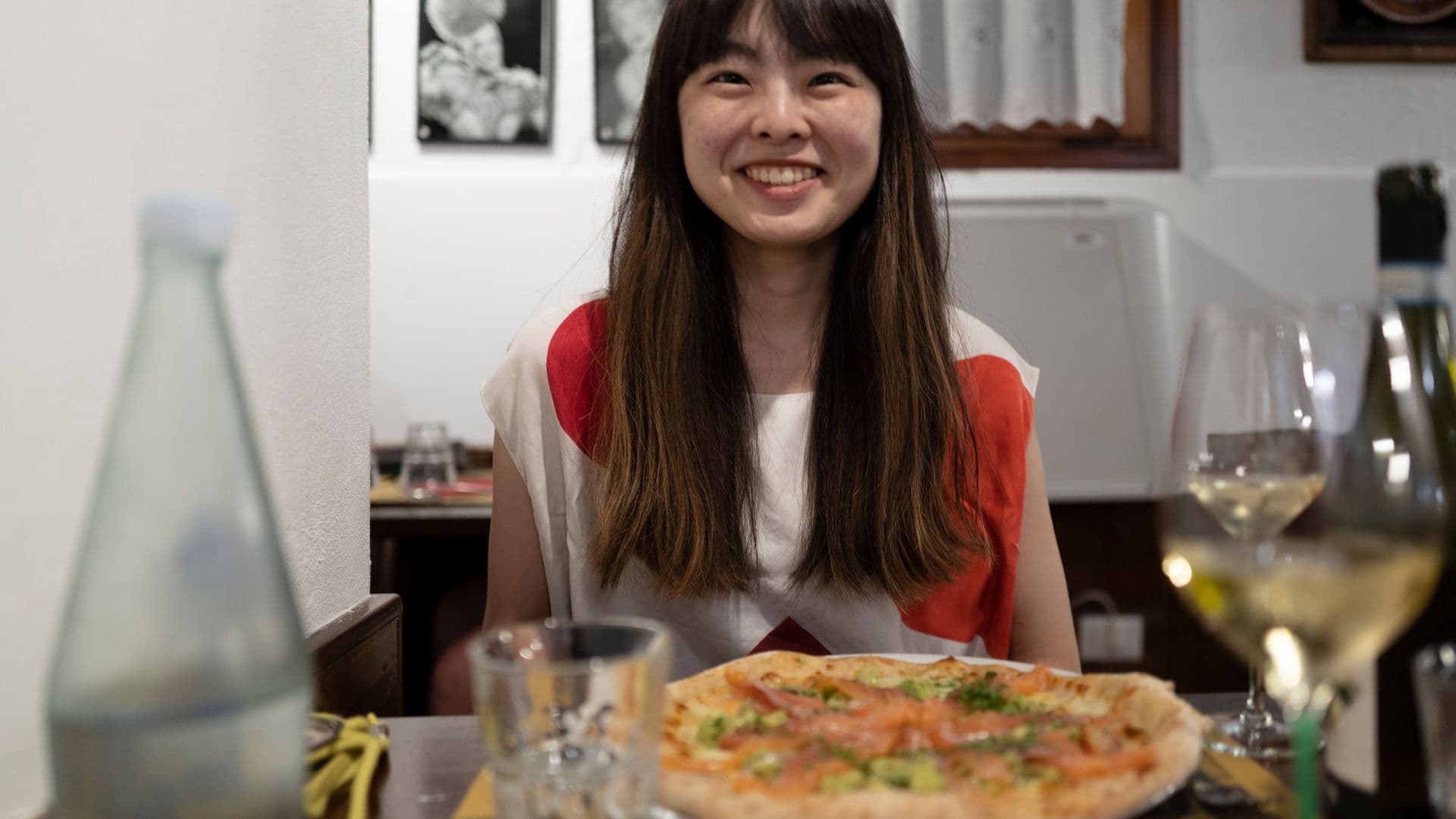 Woman smiling at a restaurant table with a pizza and a glass of white wine in front of her.