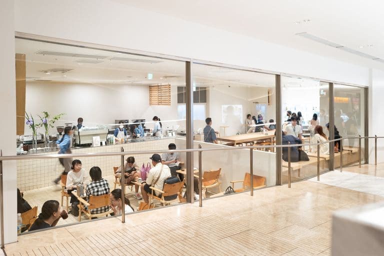 View of a modern coffee shop or cafe with customers sitting at tables and others waiting in line or ordering at the counter, with the interior visible through large glass windows.