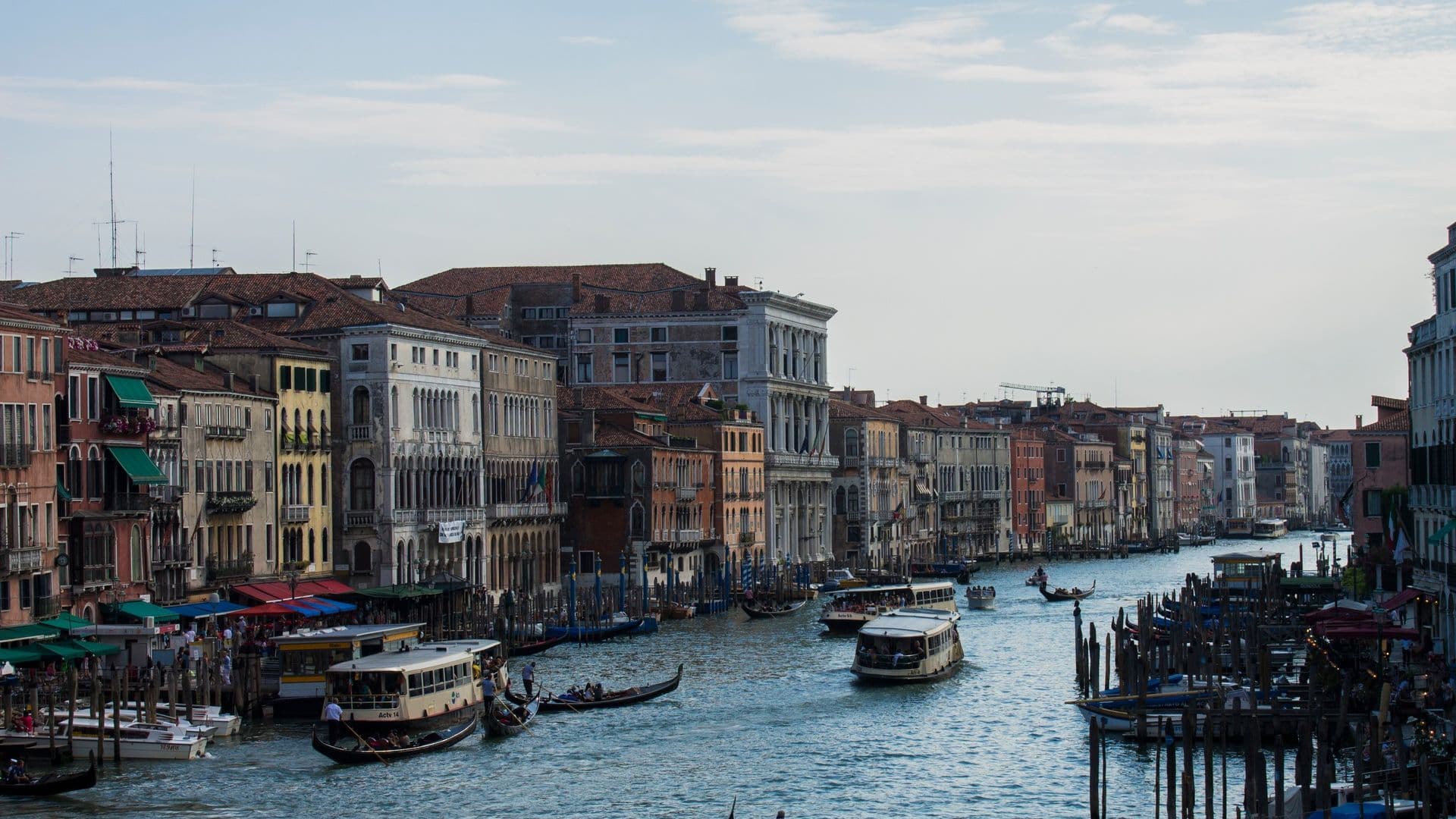 A view of the Grand Canal in Venice, Italy with historic buildings on either side, boats including gondolas and water taxis on the water, under a partly cloudy sky.