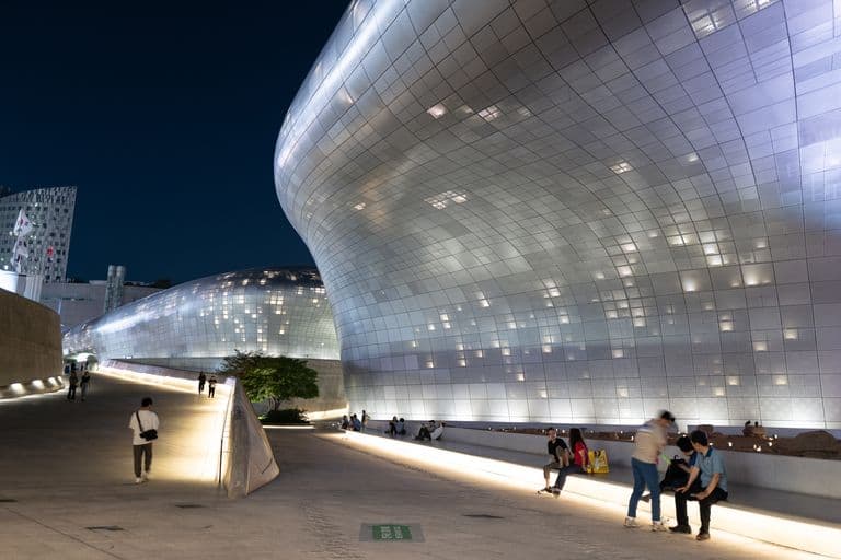 Night view of a shiny, modern architectural building with curved metallic surfaces and surrounding people sitting and walking on a lit outdoor area.