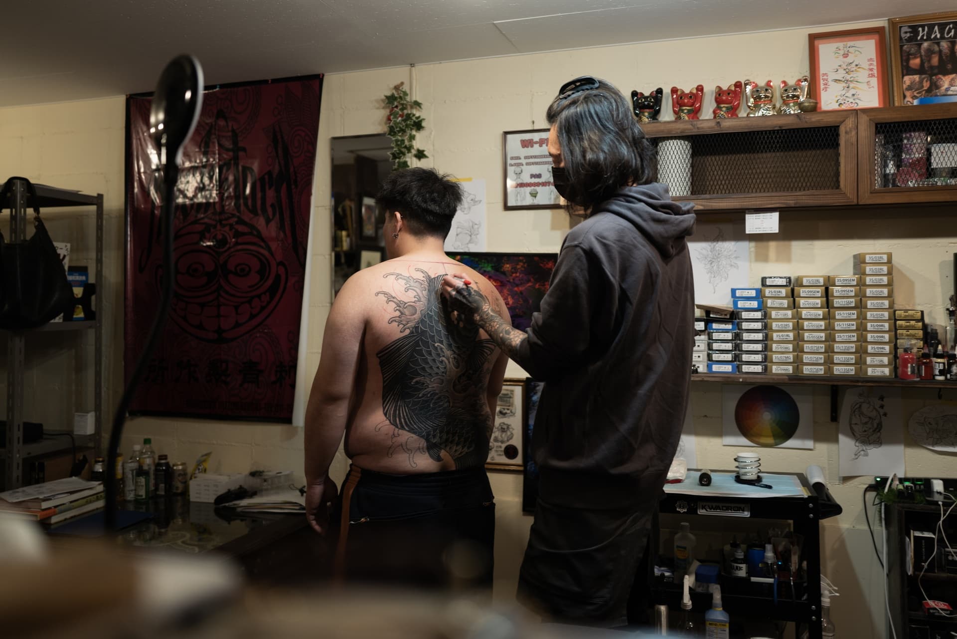 A tattoo artist is working on a large dragon tattoo on a person's back in a tattoo studio. The person has short hair and is shirtless, while the artist has long dark hair, is wearing a hoodie, and a face mask.
