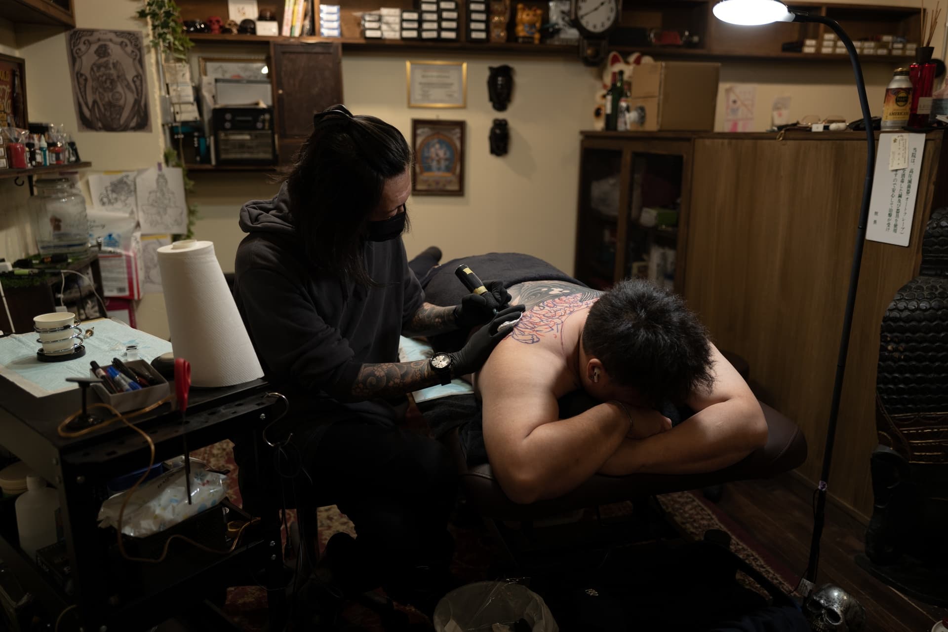 Tattoo artist in black clothing and face mask tattooing a large colorful design on a shirtless man lying on a table.