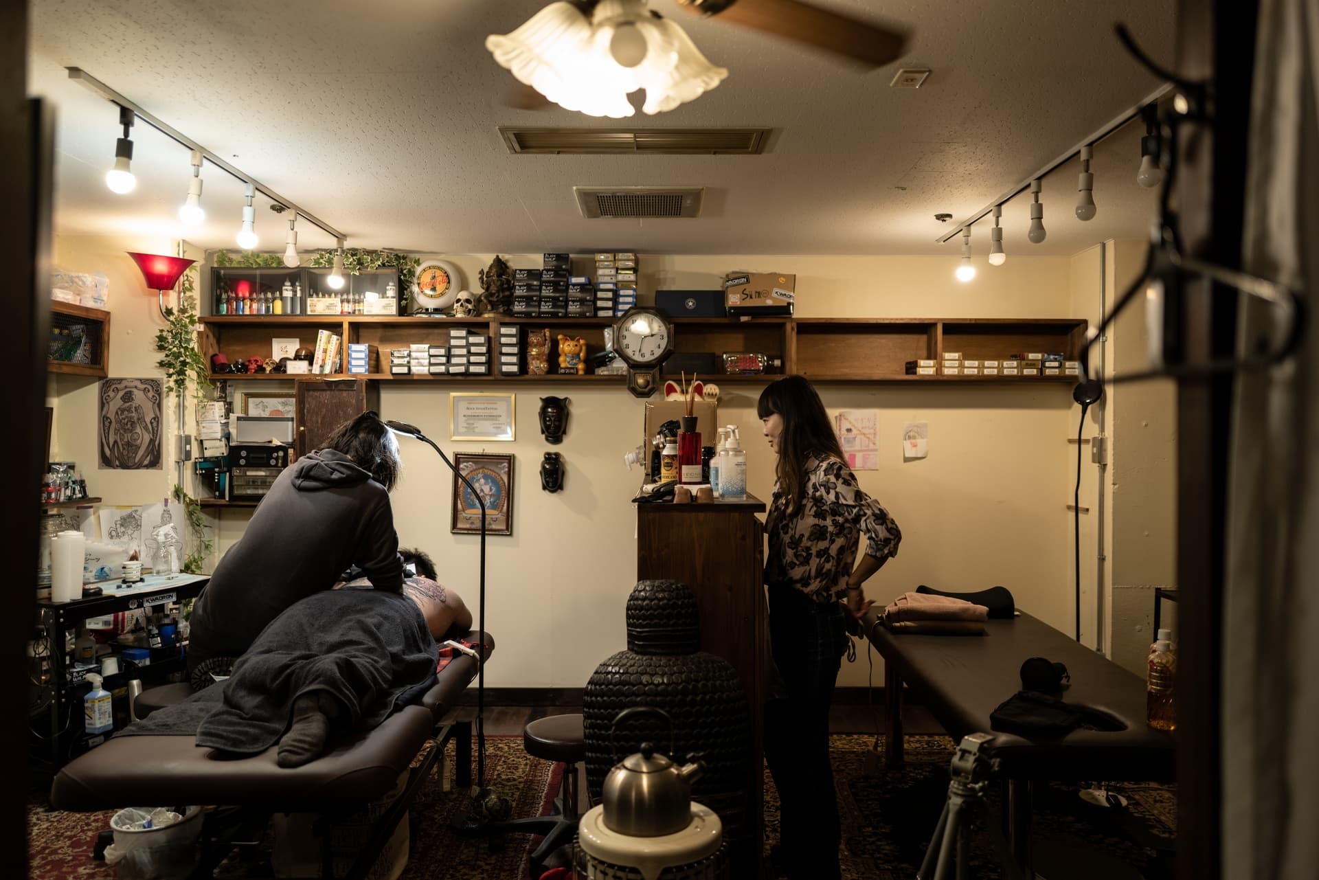 A massage therapy session taking place in a cozy room with two women, one lying face down on a massage table and the other performing the massage. A third woman stands nearby, observing. The room is decorated with shelves filled with various items, f